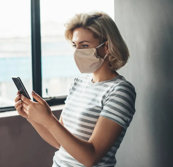 Blonda femeie cu masca medicala pe fata chat pe mobil forma acasă în timpul blocării — Fotografie, imagine de stoc