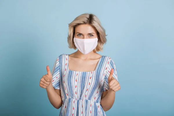 Ung kaukasisk kvinna bär en anti influensa mask och klänning samtidigt gester liknande tecken på en blå studio vägg — Stockfoto