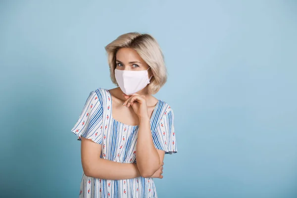 Draguta blonda doamnă în rochie și mască medicală pe față pozând pe un perete albastru cu spațiu liber — Fotografie, imagine de stoc