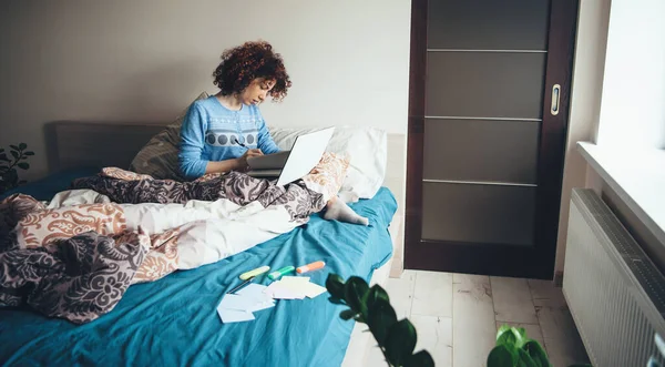 Encantadora dama con el pelo rizado usando un pijama azul está usando una computadora portátil en la cama mientras hace la tarea —  Fotos de Stock