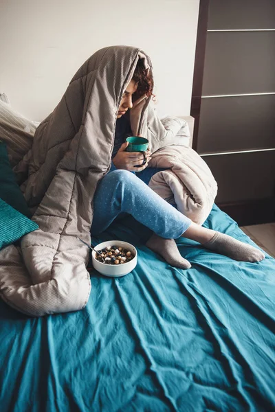 Γοητευτική κυρία με σγουρά μαλλιά που πίνει γάλα και τρώει δημητριακά στο κρεβάτι καλυμμένη με πάπλωμα — Φωτογραφία Αρχείου