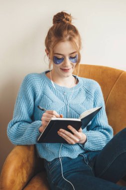 Çilli kızıl kadın kitaba bir şeyler yazıyor. Koltuğa oturmuş hidrojel göz bandı takıyor.