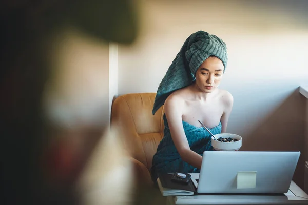 Young fitness lady sitting at the computer after shower and eating cereals with a towel on head
