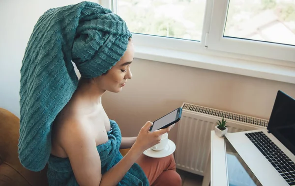 Mujer caucásica con una toalla en la cabeza está charlando en el móvil y el ordenador portátil en casa después del baño — Foto de Stock