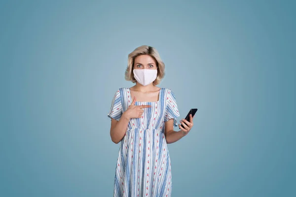Mujer caucásica con el pelo rubio y la máscara con un vestido está apuntando sorprendido por su teléfono en una pared de estudio azul — Foto de Stock