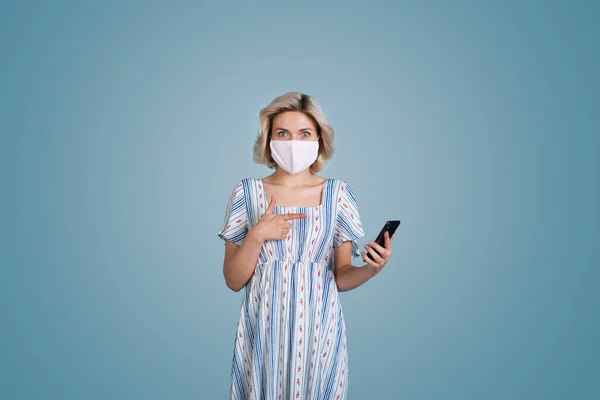 Femme caucasienne avec des cheveux blonds et masque portant une robe pointe stupéfait à son téléphone sur un mur studio bleu — Photo