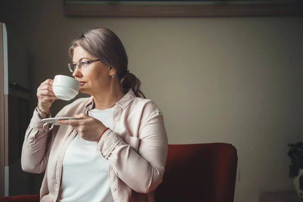 Mujer caucásica pensativa con cabello rubio bebiendo un café cerca de la ventana — Foto de Stock