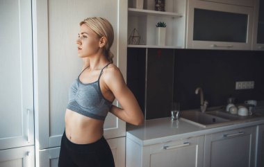 Spor giysili sarışın beyaz kadın evde fitness yapmadan önce mutfakta ısınıyor.