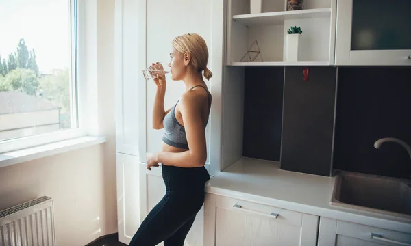 Krásná blondýna žena pije sklenici vody na sobě sportovní oblečení doma — Stock fotografie