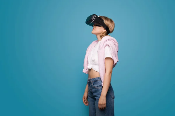 Mujer rubia joven usando auriculares de realidad virtual y posando en una pared de estudio azul — Foto de Stock