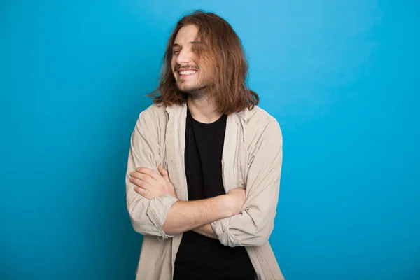 Hombre caucásico guapo con el pelo largo y la barba sonriendo con las manos cruzadas en una pared de estudio azul — Foto de Stock