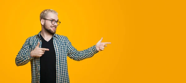 Homem caucasiano barbudo com cabelo loiro e óculos está sorrindo e apontando para o estúdio amarelo espaço livre — Fotografia de Stock