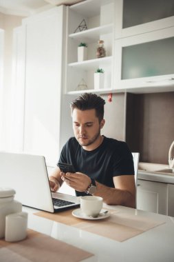 Evde laptopun başında çalışırken cep telefonuyla konuşan ve kahve içen beyaz bir adam.