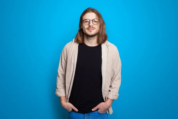 Homem elegante com cabelos longos e cerdas posando com óculos em uma parede azul do estúdio — Fotografia de Stock