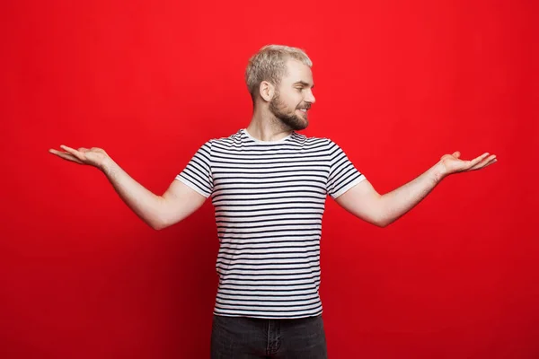 Homem loiro caucasiano com barba comparando duas coisas em suas palmas está posando em uma parede de estúdio vermelho — Fotografia de Stock