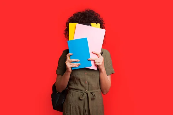 Estudiante caucásica con el pelo rizado cubriendo su cara con libros y posando en una pared de estudio rojo sosteniendo una bolsa — Foto de Stock