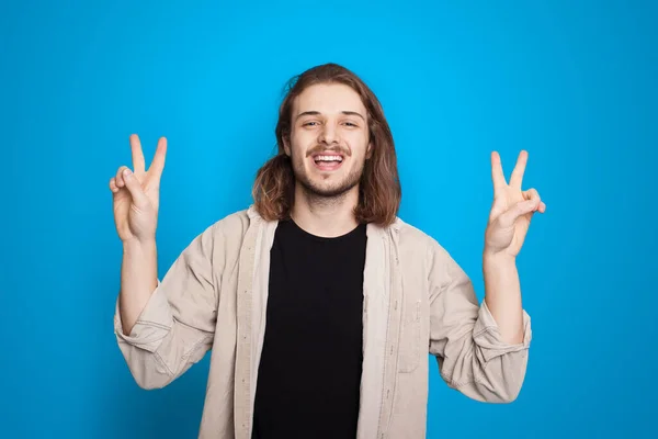 Cáucaso homem com cabelos longos gesticulando o sinal de olá e peça está sorrindo em um fundo de estúdio azul — Fotografia de Stock