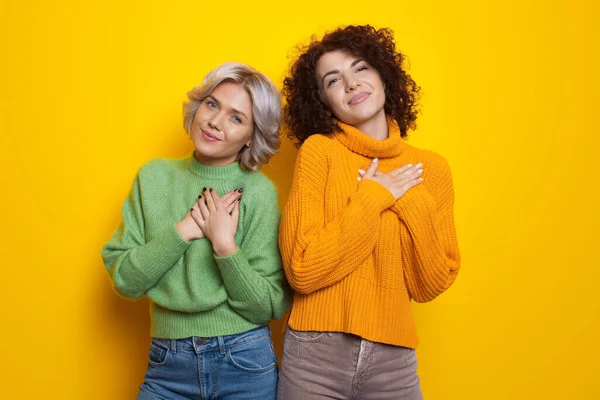 Çekici kıvırcık saçlı kız kardeşler, sarı bir stüdyo duvarında aşk işareti yapan avuç içleriyle kalplerine dokunuyor. — Stok fotoğraf