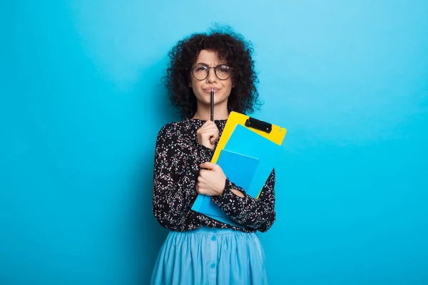 Nachdenkliche kaukasische Studentin mit lockigem Haar posiert mit einem Buch in einem Kleid an einer blauen Studiowand — Stockfoto