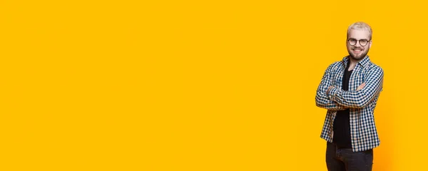 Homem loiro caucasiano anunciando algo em uma parede de estúdio amarelo enquanto usava óculos e posando com as mãos cruzadas perto do espaço livre — Fotografia de Stock