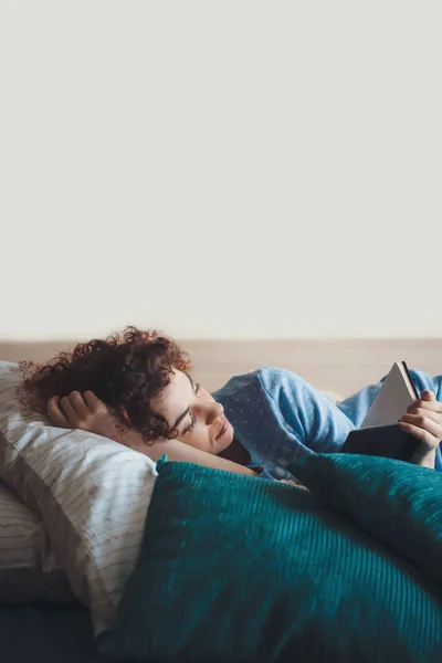 Mujer caucásica con hábitos saludables y cabello rizado usando un pijama azul en la cama leyendo un libro acostado en la almohada —  Fotos de Stock
