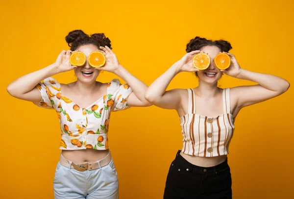 Vorderseite Foto von zwei kaukasischen Zwillingen, die ihr Auge mit Orangen bedecken und an einer gelben Studiowand lächeln — Stockfoto