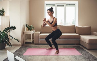 Spor kıyafetli bir kadın dizüstü bilgisayar kullanıyor ve evdeki yoga halısında bacaklarını çalıştırıyor.