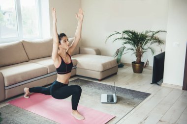 Esneyen beyaz kadın spor kıyafetlerini giyip evde spor yapıyor ve yerde dizüstü bilgisayar kullanıyor.