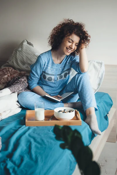 Foto vista frontal de una mujer de pelo rizado sonriendo en un pijama azul mientras lee un libro en la cama y come cereales con leche — Foto de Stock