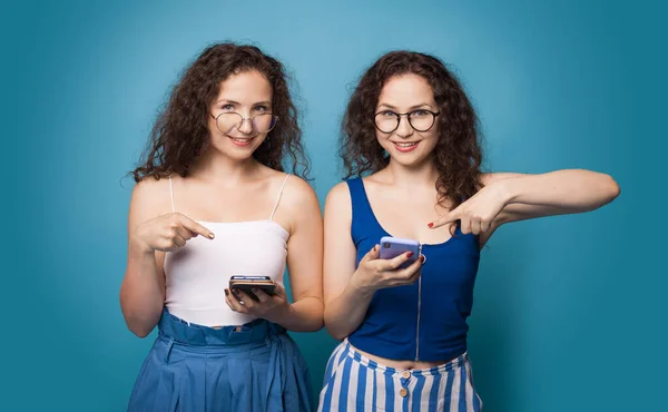 Zwei kaukasische Schwestern, die auf das Telefon zeigen, während sie eine Brille und lockiges Haar an einer blauen Studiowand tragen — Stockfoto