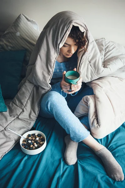 Foto vista superior de una mujer de pelo rizado bebiendo una taza de leche mientras come cereales en la cama cubierta con una colcha — Foto de Stock