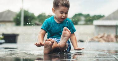 Küçük beyaz bir çocuk dışarıda, yağmurun altında mavi bir tişört giyerek oynuyor.