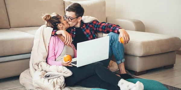 Υπέροχο ζευγάρι Καυκάσιος κάθεται στο πάτωμα και φιλιά, ενώ χρησιμοποιώντας ένα φορητό υπολογιστή και κρατώντας ένα πορτοκάλι που καλύπτεται με ένα πάπλωμα — Φωτογραφία Αρχείου