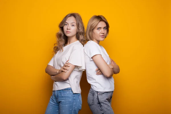 Nette blonde Frauen posieren Rücken an Rücken auf einer gelben Studiowand mit gekreuzten Händen — Stockfoto
