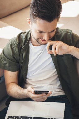 Gülümseyen beyaz adam cep telefonunda sohbet ediyor ve dizlerinin üstünde dizüstü bilgisayar tutuyor.