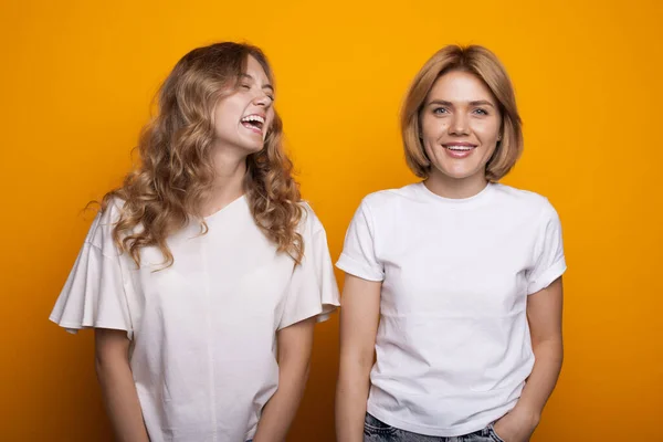 Lächelnde blonde Frau mit lockigem Haar posiert neben ihrer Schwester auf einer gelben Studiowand in einem weißen T-Shirt — Stockfoto