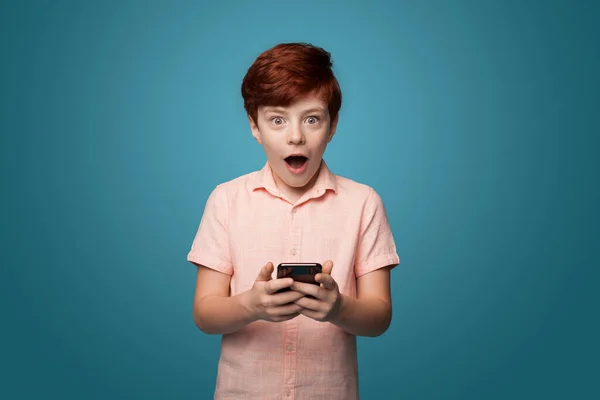 Chocado caucasina gengibre menino segurando um telefone e olhando para a câmera em uma parede de estúdio azul — Fotografia de Stock