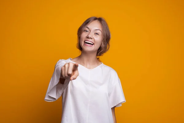 Femeia atractivă zâmbind și arătând spre camera foto pe un perete galben de studio purtând un tricou alb — Fotografie, imagine de stoc