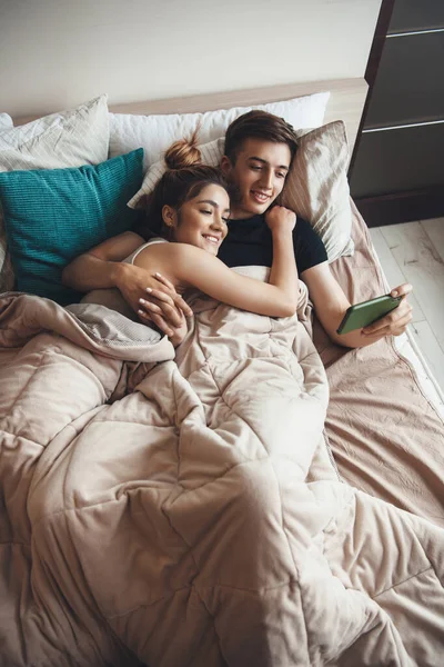 Vue supérieure photo d'un couple caucasien couché dans le lit le matin et regardant quelque chose sur mobile tout en embrassant et sourire — Photo