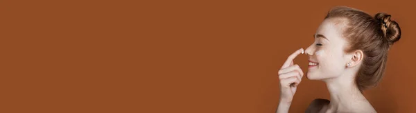 Рыжая женщина с веснушками наносит крем на лицо и улыбается голыми плечами на коричневую стену студии с свободным пространством — стоковое фото