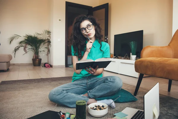 Mujer caucásica de pelo rizado con gafas que comen cereales con jugo de verduras frescas está haciendo la tarea en el suelo usando un libro y un ordenador portátil — Foto de Stock