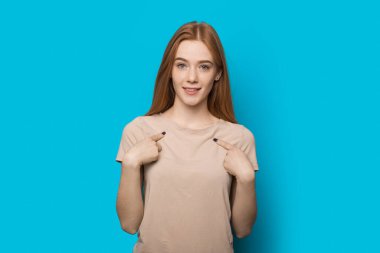 Confident caucasian woman with red hair and freckles posing on a blue wall is pointing to her and look at camera clipart