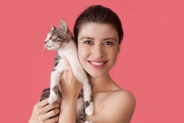 Çilli, beyaz, çıplak omuzlu, pembe duvarda poz veren, kameraya gülümseyen kedisi olan bir kadın.