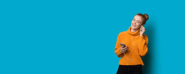 Mulher caucasiana alegre com sardas e cabelos vermelhos está ouvindo música usando fones de ouvido e celular em uma parede azul com espaço em branco — Fotografia de Stock