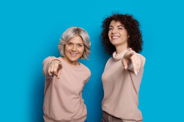Dos damas caucásicas con el pelo rizado apuntan a la cámara y sonríen felices en una pared azul — Foto de Stock