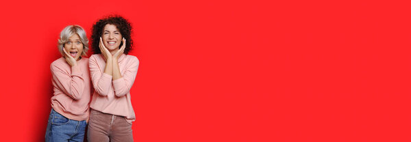 Two surprised caucasian women in pink sweaters and curly hair are posing on a red wall with free space