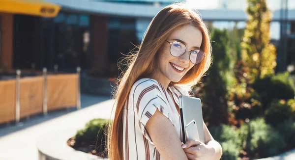 Mujer caucásica pecosa con el pelo rojo y anteojos abrazando un ordenador portátil y posando en la cámara con un teléfono — Foto de Stock