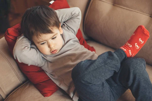 Upper side view of a caucasian boy looking down and feeling sad while lying on a sofa with a red pillow