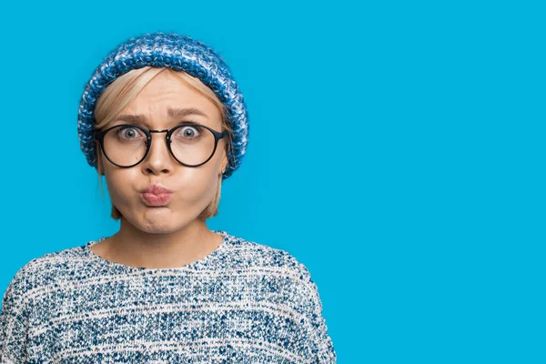 Deliciosa mujer caucásica con el pelo rubio con sombrero de punto y suéter está mirando a través de las gafas y haciendo la cara divertida en la cámara en una pared azul — Foto de Stock
