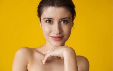 Kahverengi saçlı ve çilli neşeli beyaz kadın kameraya bakıyor çenesine dokunuyor ve sarı bir duvarda çıplak omuzlarıyla poz veriyor.